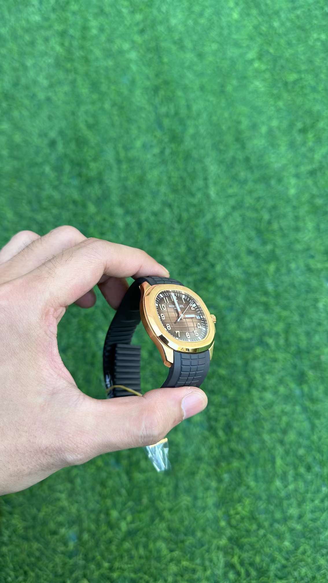 Hand-held distant shot of Patek Philippe Aquanaut 5167R-001 showing full watch silhouette on green turf.