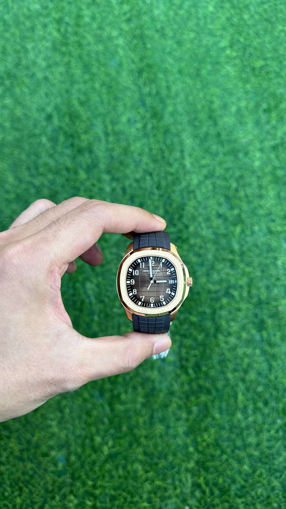 Outdoor hand-held shot of Patek Philippe Aquanaut 5167R-001 showing dial and rose-gold bezel on green background.